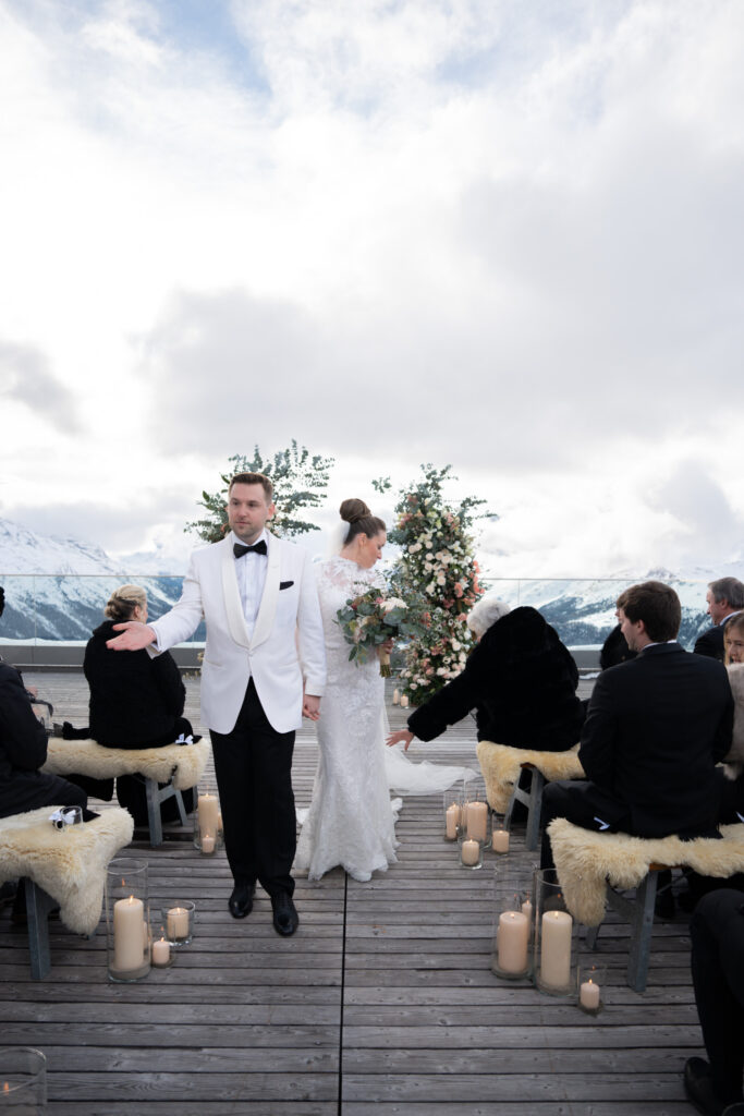 Eine Braut und ein Bräutigam stehen bei einer Hochzeitszeremonie im Freien, die Gäste sitzen und im Hintergrund sind verschneite Berge zu sehen. Perfekt eingefangen von einer Hochzeitsfotografin, die sich auf Destination Weddings spezialisiert hat.