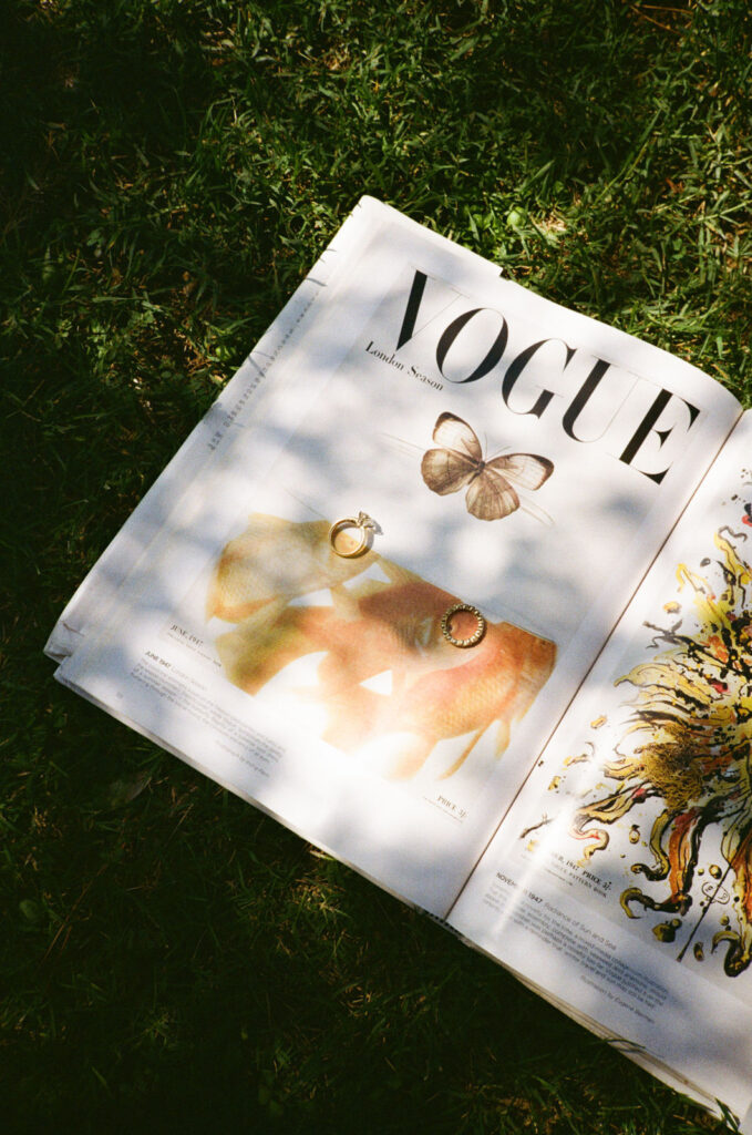Ein aufgeschlagenes Vogue-Magazin liegt im Gras, Sonnenlicht und Schatten fallen auf das Titelblatt mit Schmuck und einem Schmetterling.