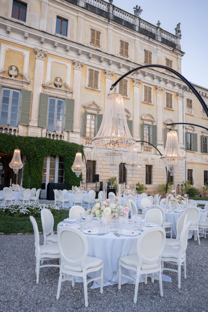 Elegante Hochzeitsfeier im Freien mit runden Tischen, weißen Stühlen und Kronleuchtern vor einer großen Villa.