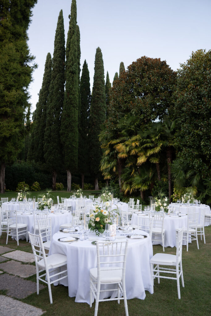 Elegant al fresco dining with white-covered tables and chairs, surrounded by tall trees and lush greenery.