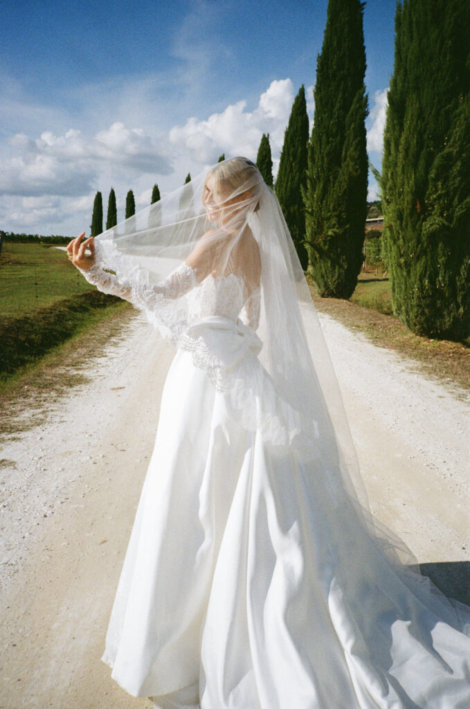 Eine Braut in weißem Kleid und Schleier geht eine von Bäumen gesäumte Landstraße unter einem blauen Wolkenhimmel entlang.