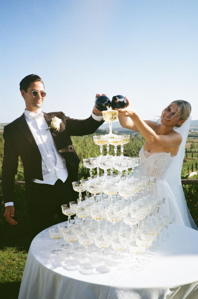 Braut und Bräutigam gießen an ihrem Hochzeitstag im Freien Sekt in einen hohen Turm aus Gläsern.