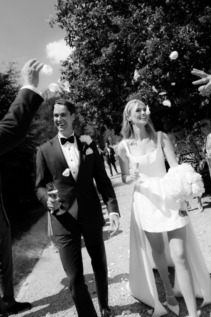 The bride and groom smile as they walk outdoors and guests throw petals at their wedding.