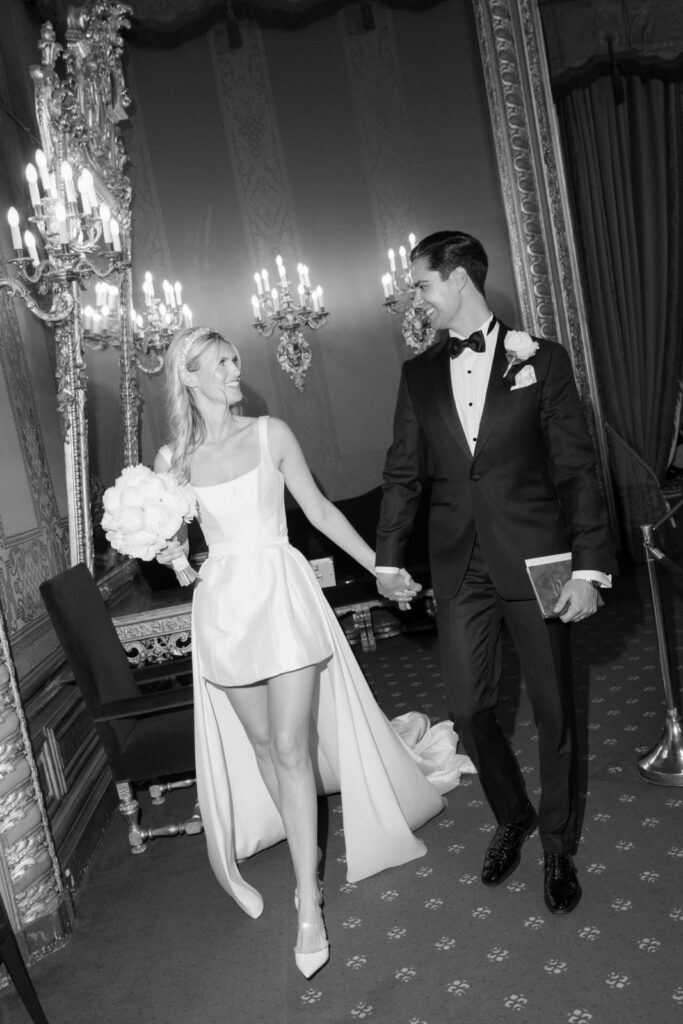 A bride and groom hold hands and smile at each other in an elegant, ornate room.