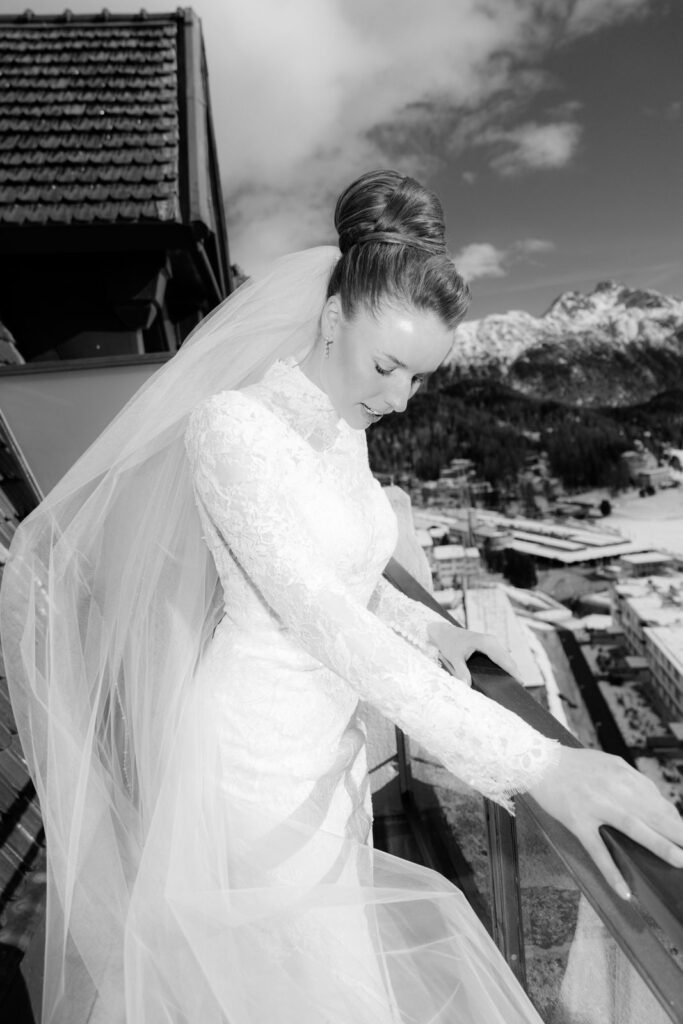 The bride in a lace wedding dress with a veil stands on a balcony with snow-capped mountains in the background.