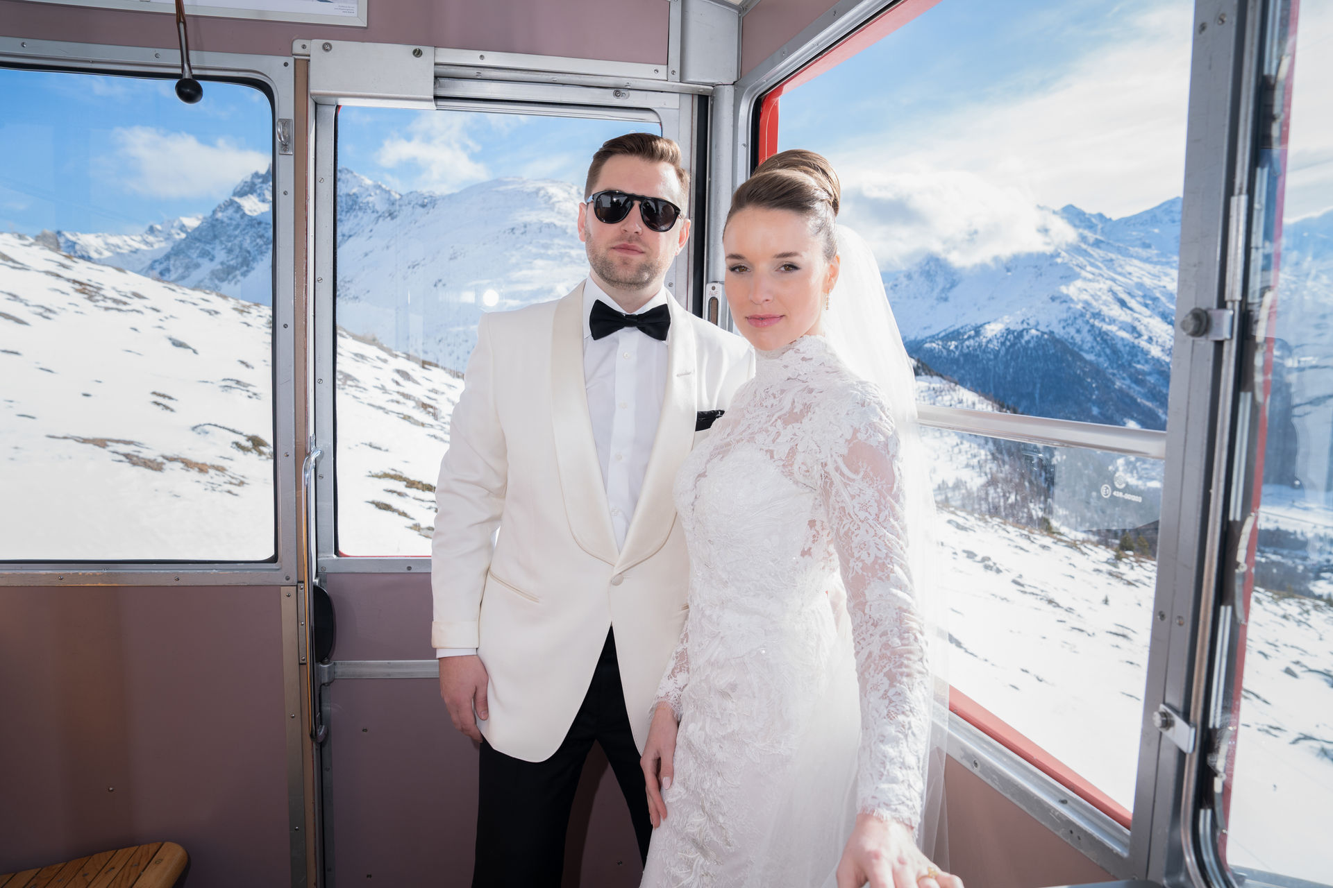 Bride and groom in formal attire posing in a cable car with snow-covered mountains in the background.