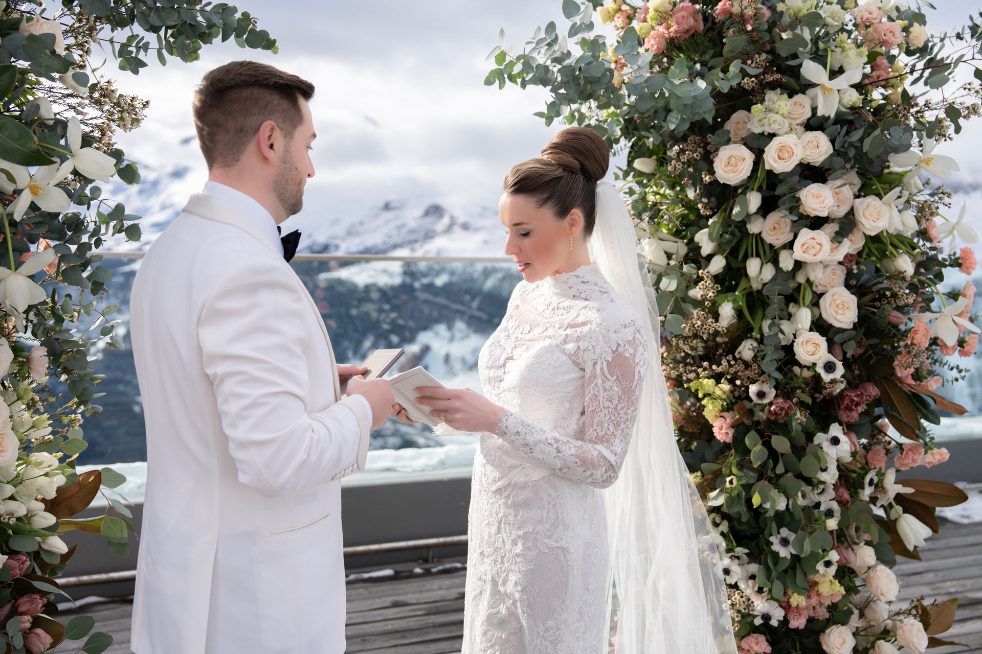 Wedding photographer Austria A bride and groom exchange their wedding vows outdoors under floral arches, with snow-capped mountains in the background.