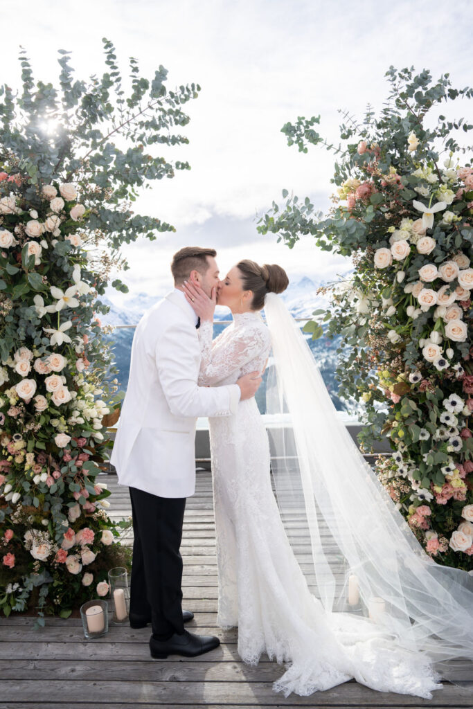 Bride and groom kissing at an outdoor wedding altar framed by flowers, mountains in the background.
