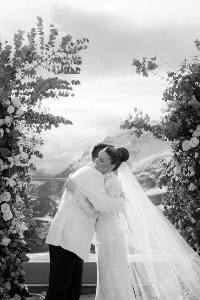 Braut und Bräutigam umarmen sich im Freien, eingerahmt von Blumenbögen, mit Bergen im Hintergrund.