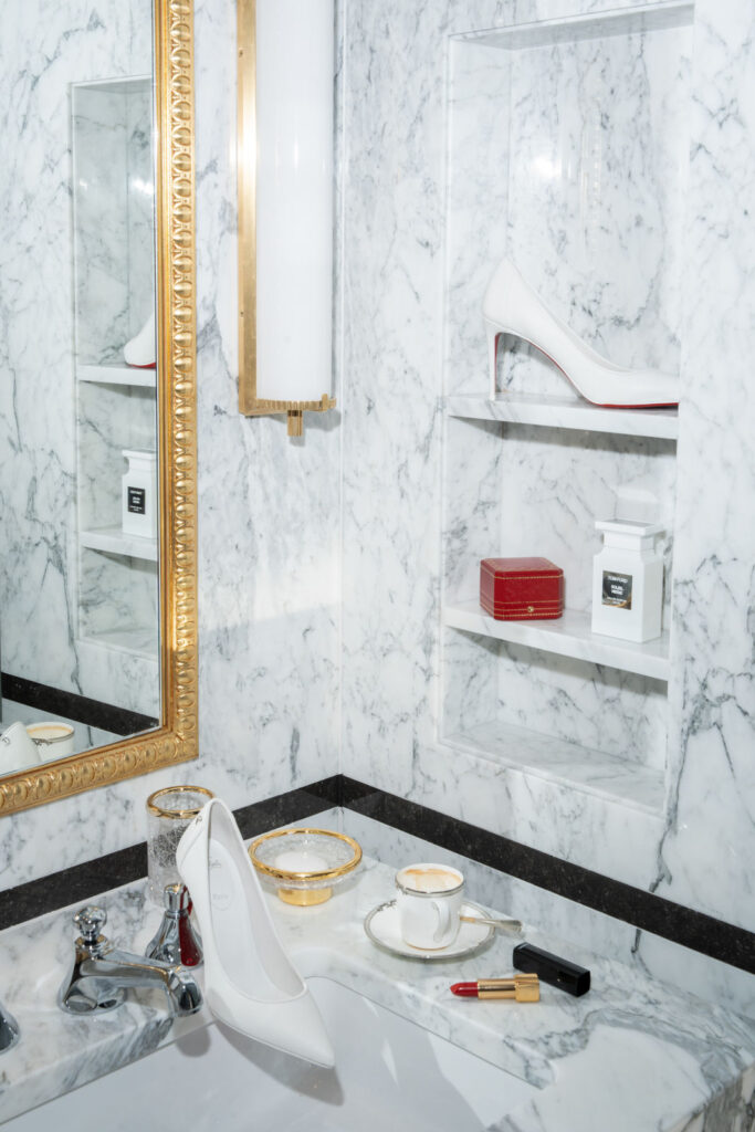 Weiße High Heels, Lippenstift, Teetassen und Parfüm auf einem Marmorwaschbecken mit goldenem Spiegel und Regalen.