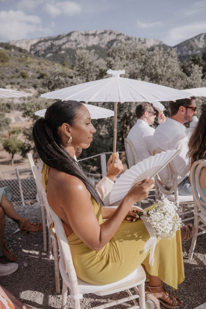 Eine Frau in einem gelben Kleid hält bei einer Hochzeit in Son Marroig einen weißen Sonnenschirm und einen Handfächer in der Hand.