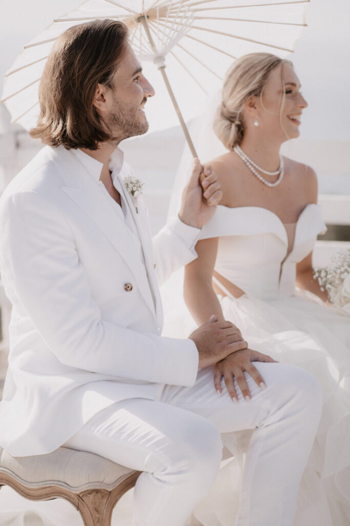Ein Brautpaar in weißer Hochzeitskleidung, das gemeinsam unter einem Sonnenschirm in Son Marroig sitzt.