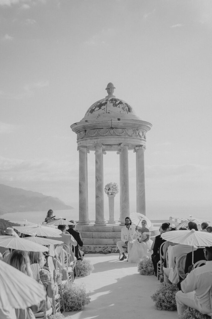 Wedding in Son Marroig: Getting married over the cliffs of Mallorca, with experiences, costs and tips 22 A wedding couple sits under a stone rotunda in Son Marroig, while the guests stand with umbrellas nearby.