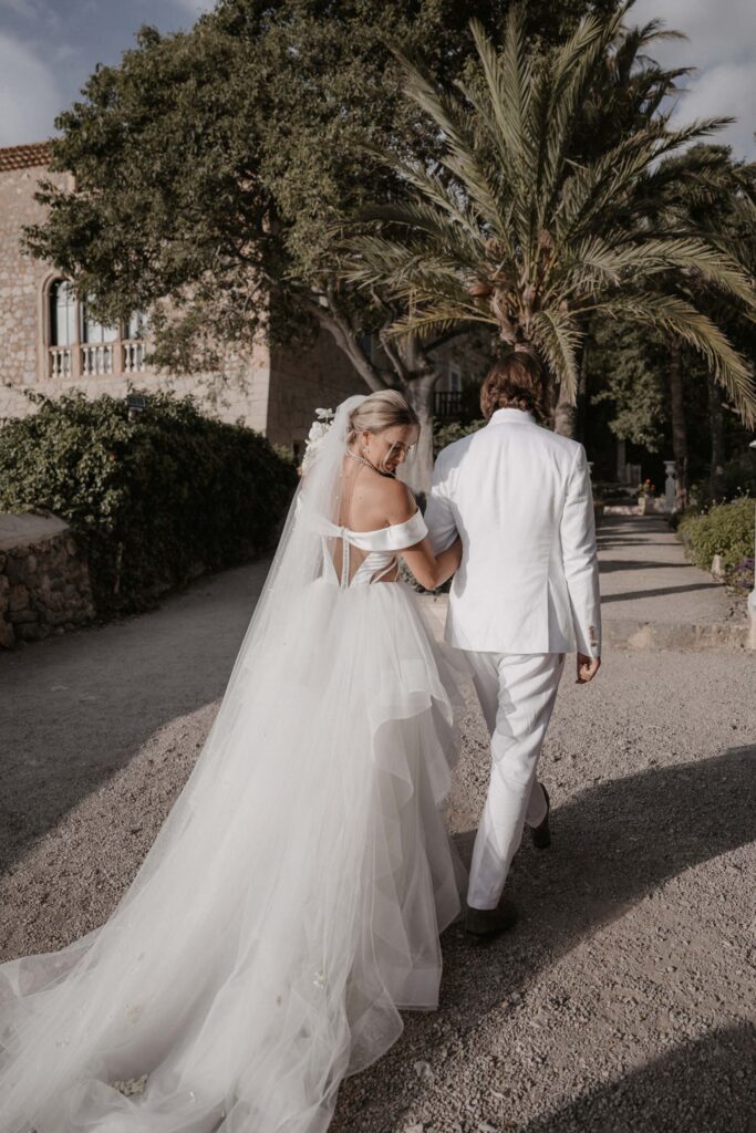 Ein Brautpaar in weißer Hochzeitskleidung, das gemeinsam auf einem Schotterweg in Son Marroig spaziert.