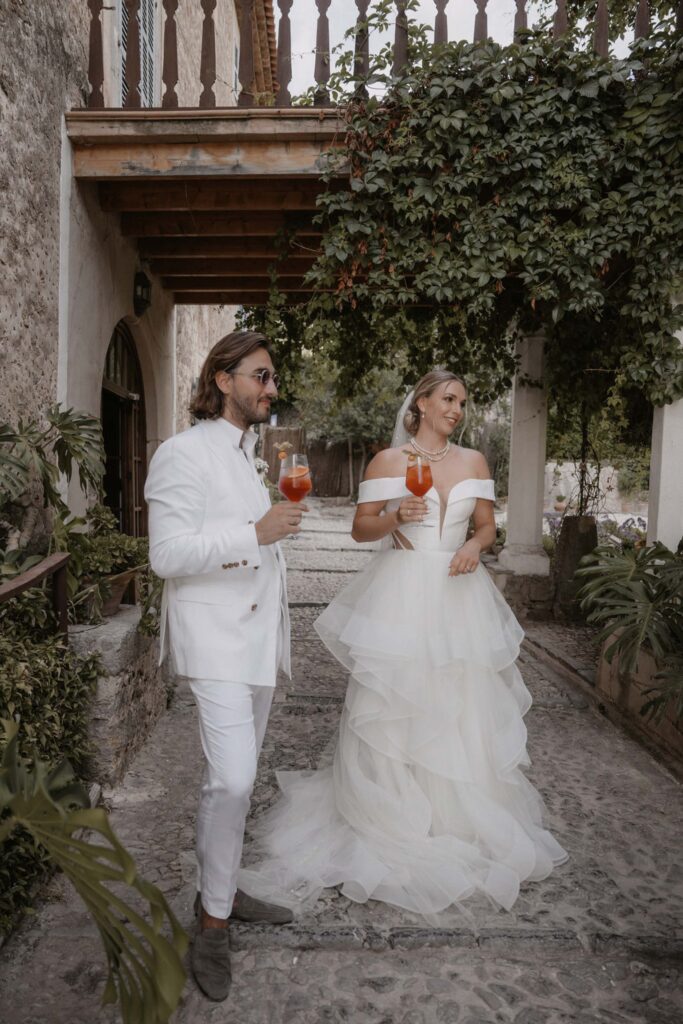 Ein Brautpaar in Hochzeitskleidung, das auf einem Steinweg bei Son Marroig Cocktails in der Hand hält.
