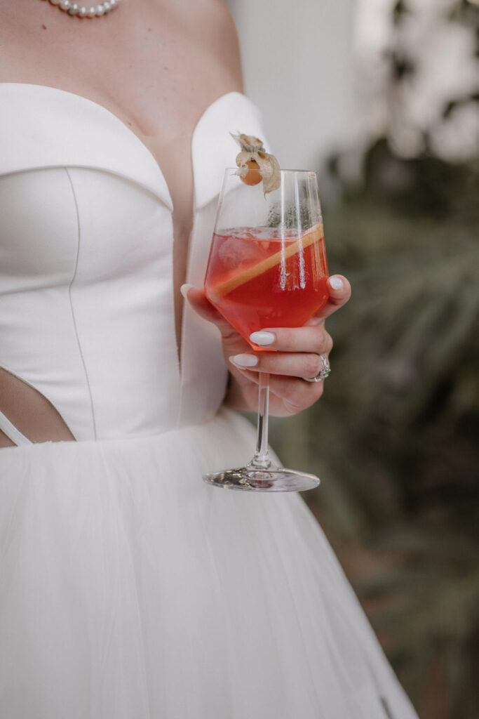 Eine Braut in Son Marroig hält einen roten Cocktail in der Hand, der mit einer Orangenscheibe und einer Physalis garniert ist.