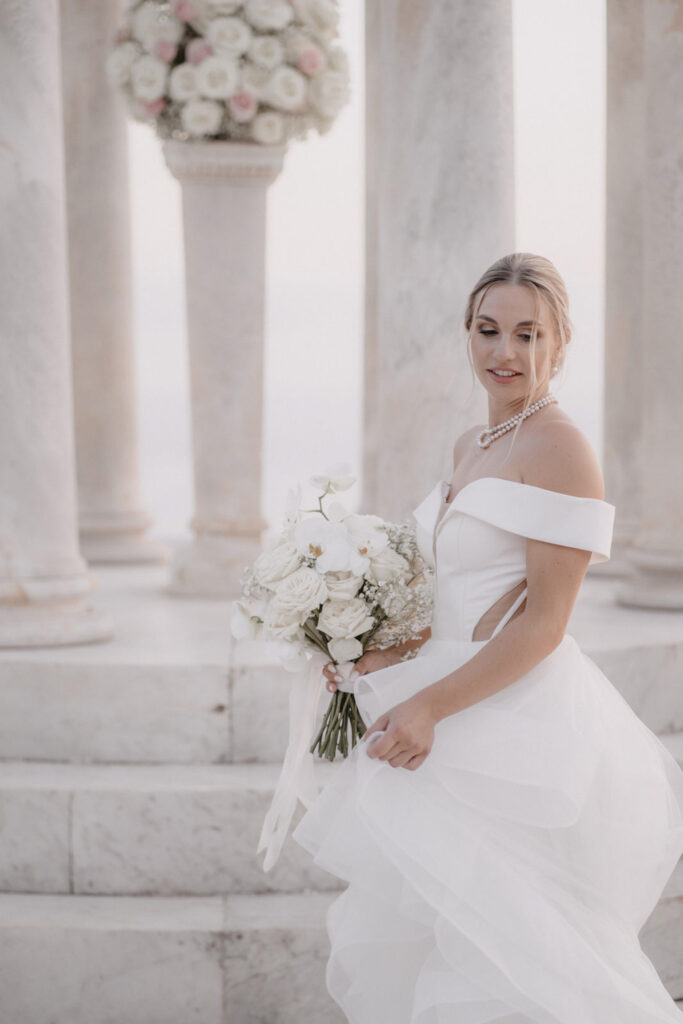 Eine Braut in einem schulterfreien Kleid hält vor den Steinsäulen von Son Marroig einen weißen Blumenstrauß in der Hand.