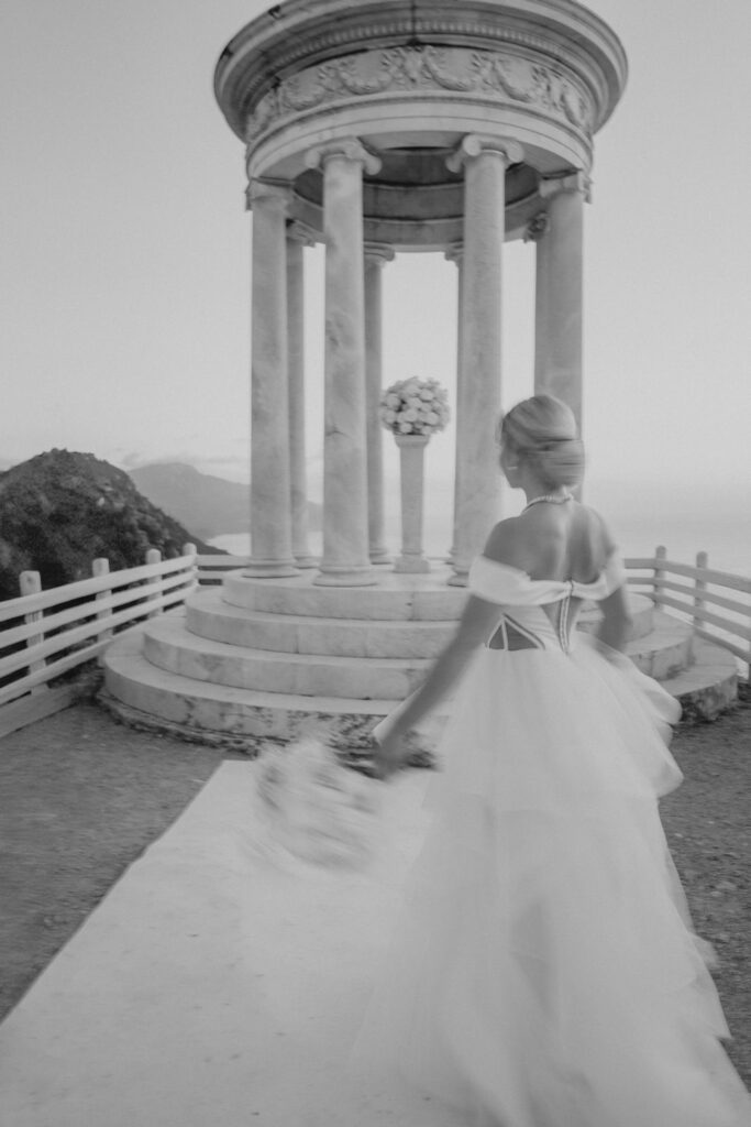 Eine Braut in einem weißen Brautkleid geht auf einen steinernen Pavillon in Son Marroig zu, von dem aus man einen Blick auf das Meer hat.