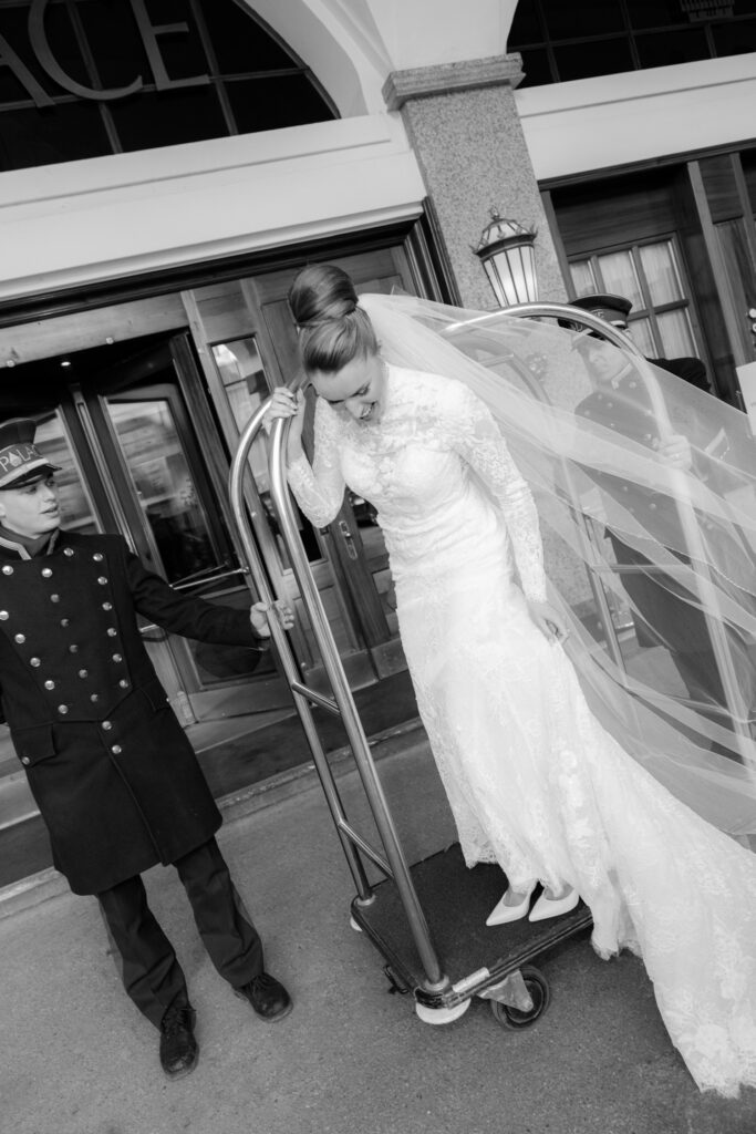A bride in a wedding dress stands on a luggage trolley with a hotel employee helping her - perfect for destination weddings, shot by a wedding photographer.