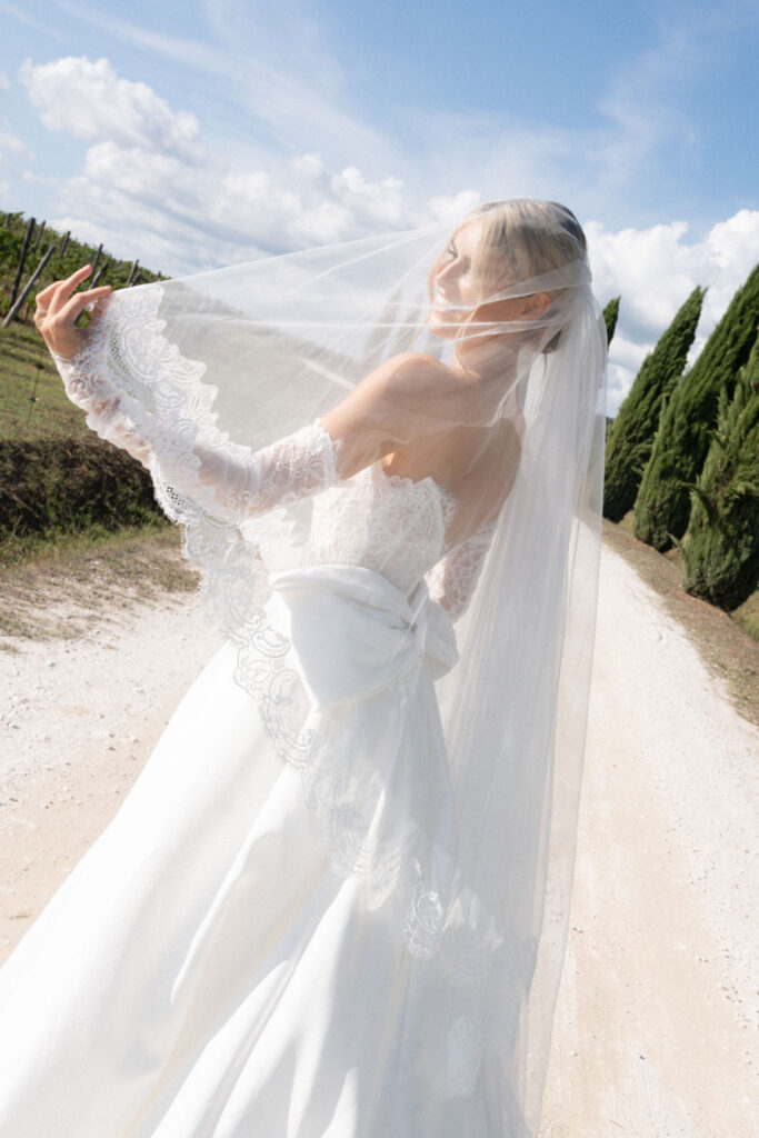 A bride in a white wedding dress and veil stands on a sunny country road, looking over her shoulder – the perfect inspiration for destination wedding photography and unforgettable elopements.