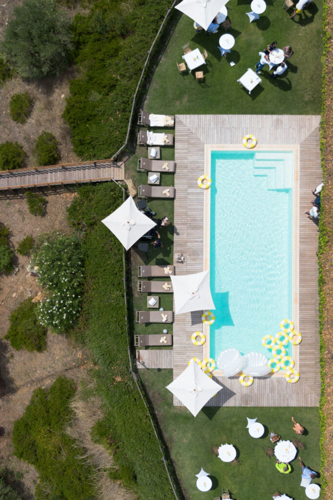 Aerial view of a pool with yellow inflatables, sun loungers, parasols and people sitting at tables on the lawn – an idyllic setting perfect for destination weddings or breathtaking overseas wedding photography.