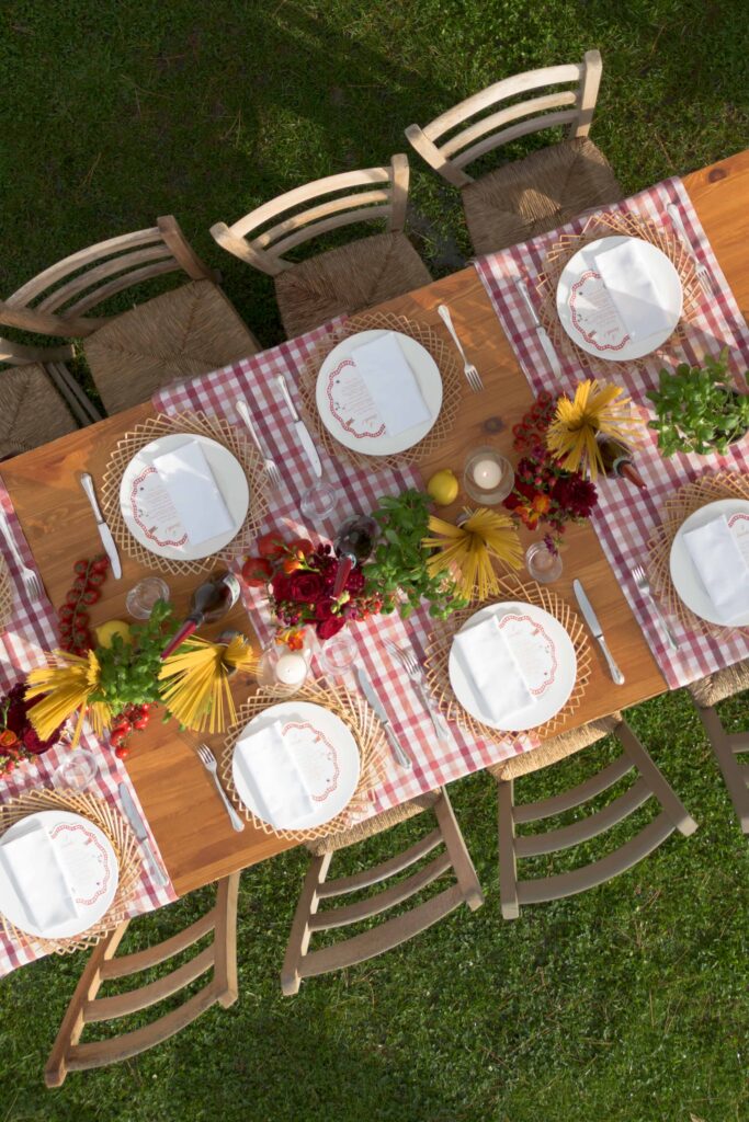 A wooden outdoor table set for a meal, with red flowers, checked napkins, and empty plates on the chairs - the perfect inspiration for destination weddings or wedding photographers looking for unique backdrops.