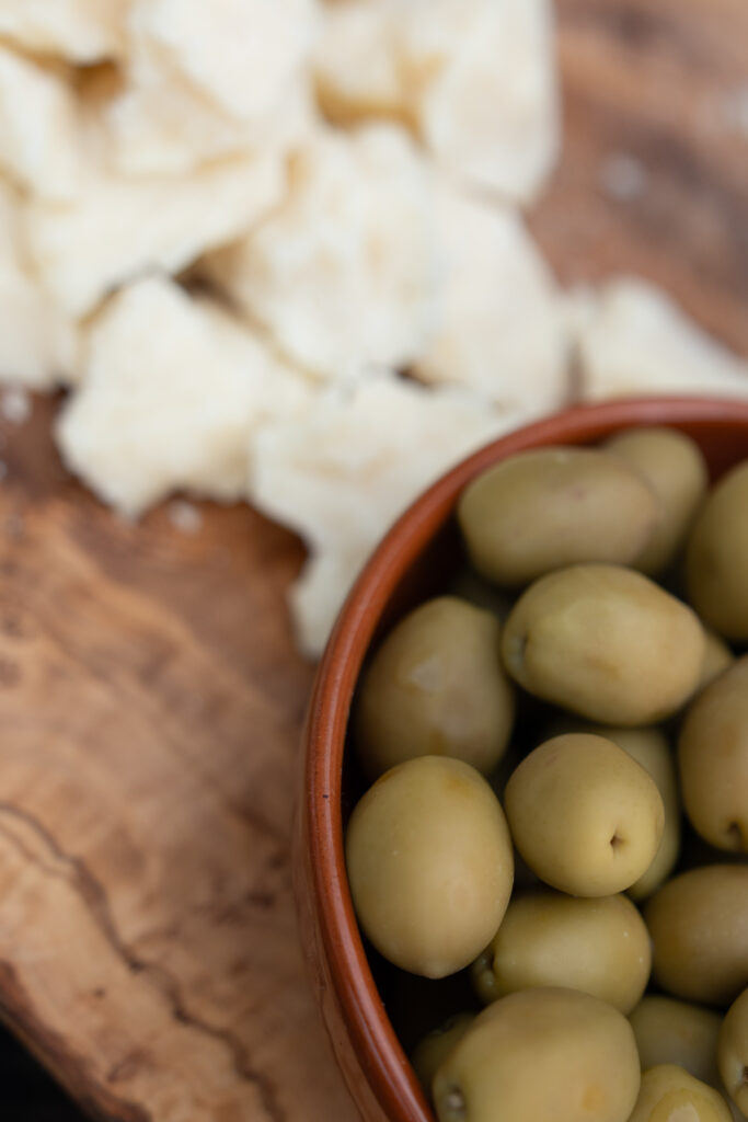 Green olives in a brown bowl next to white cheese pieces on a wooden board - perfect for sharing at your destination wedding or as inspiration for creative wedding photography.