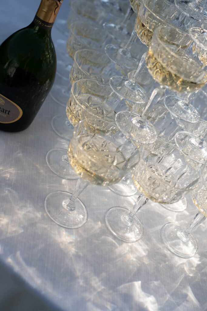 A bottle of champagne next to stacked, filled champagne flutes on a sun-drenched, white tablecloth - perfect for overseas wedding photography and capturing stunning destination weddings.