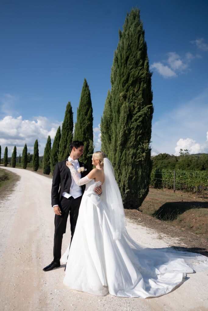 Conti di San Bonifacio Wine Resort: Exklusive Hochzeit in der Toskana, Italien 74 Eine Braut und ein Bräutigam stehen auf einem Feldweg neben hohen Bäumen und lächeln sich an ihrem Hochzeitstag an, wunderschön eingefangen von Hochzeitsfotografin, spezialisiert auf Hochzeitsfotografie im Ausland.