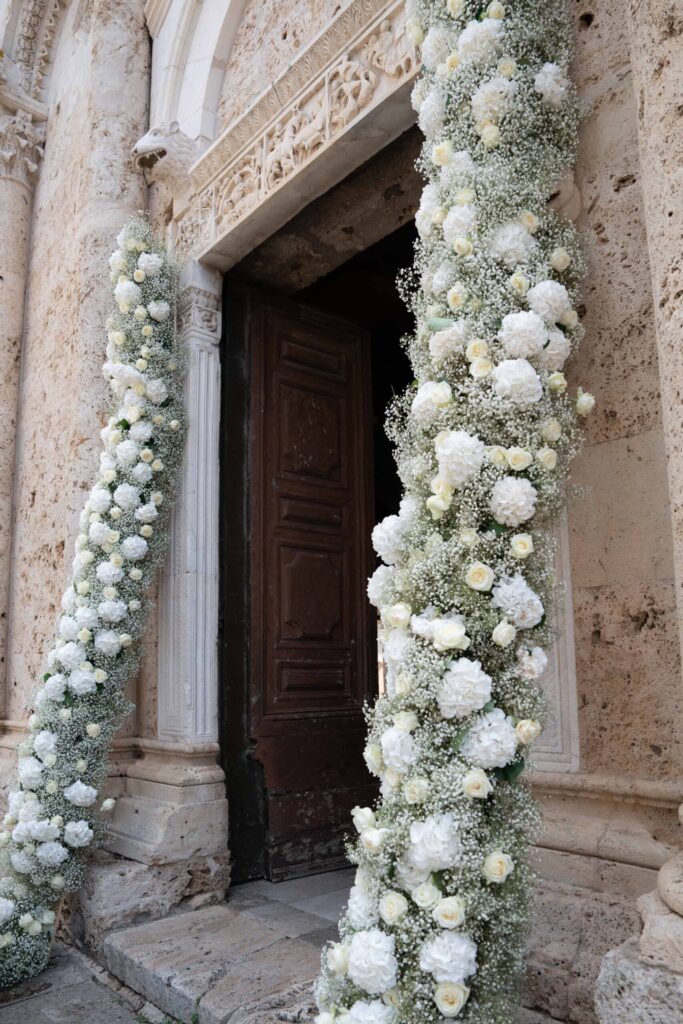 Conti di San Bonifacio Wine Resort: Exklusive Hochzeit in der Toskana, Italien 76 Zwei Säulen neben einem steinernen Portal sind mit weißen Blumen und Grünpflanzen geschmückt und bilden eine perfekte Kulisse für Hochzeiten und exquisite Hochzeitsfotos.