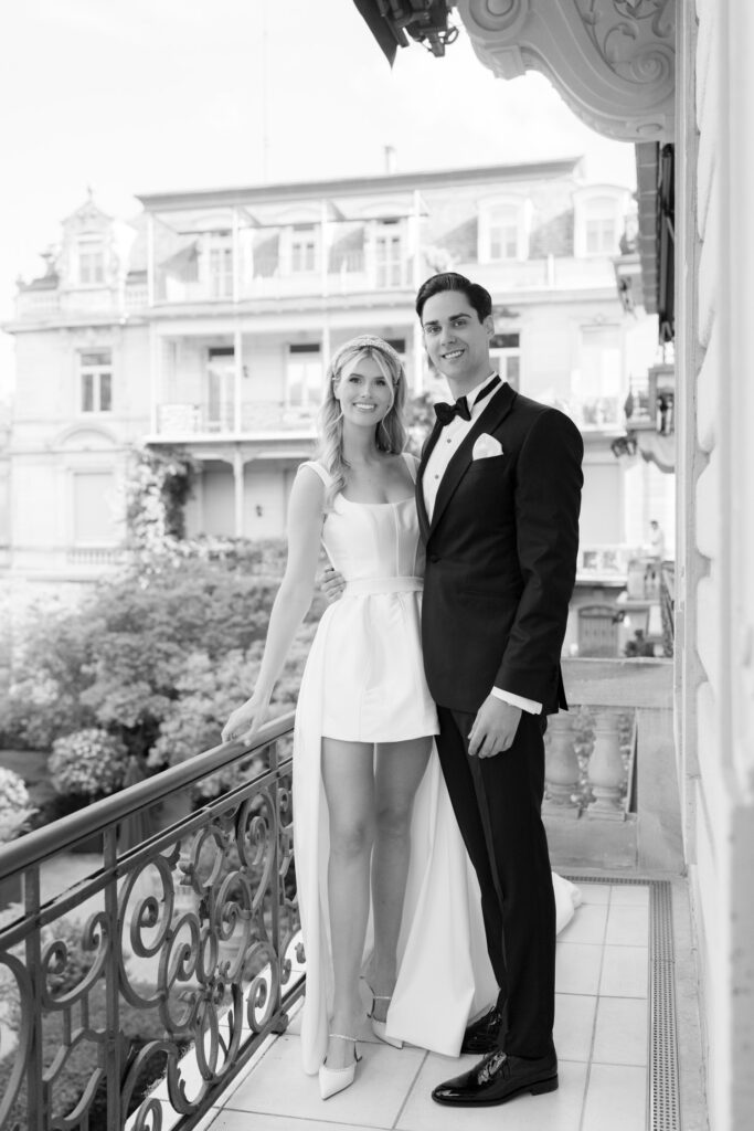 Ein Paar in formeller Kleidung posiert und lächelt auf einem Balkon mit Blick auf elegante Gebäude, in Schwarz und Weiß - aufgenommen von einer Hochzeitsfotografin, die sich auf Destination Weddings und Auslandshochzeit-Momente spezialisiert hat.
