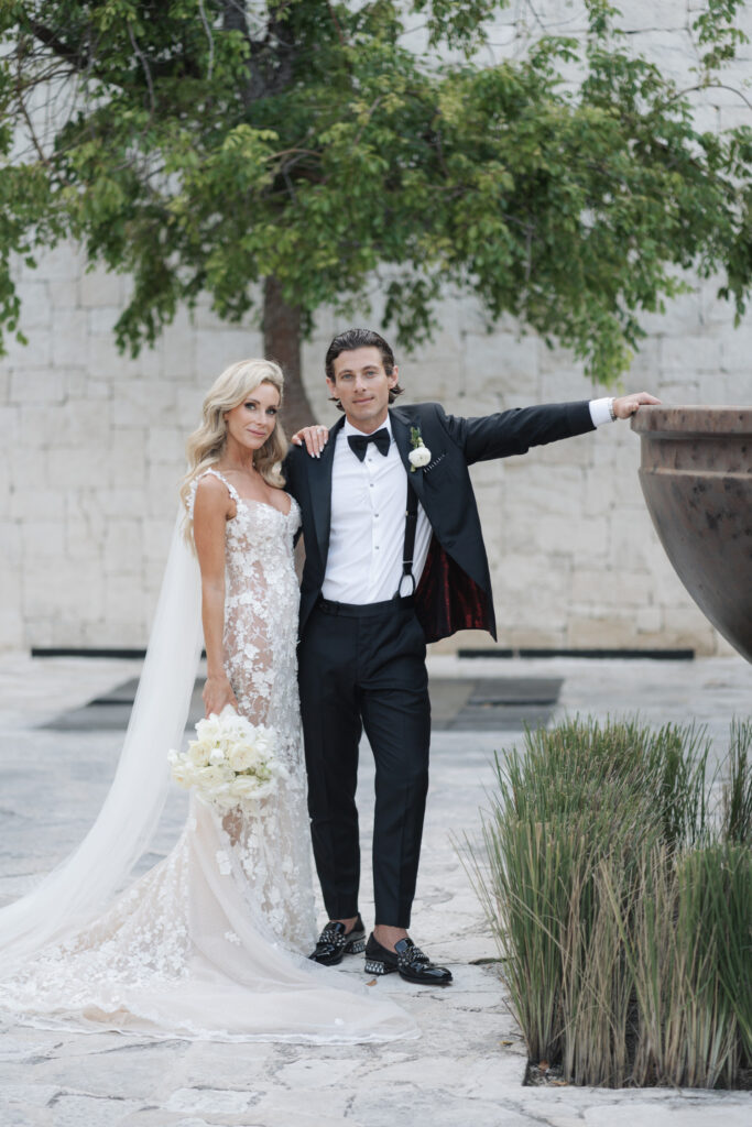 Editorial Wedding Photography: The Trend for Stylish Wedding Photos A bride in a lace dress and a groom in a tuxedo pose together outdoors in front of a tree and a stone wall, beautifully captured by a wedding photographer who specialises in wedding photography.