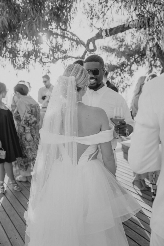 Wedding in Son Marroig: Getting married over the cliffs of Mallorca, with experiences, costs and tips 25 A bride in a white dress stands opposite a man wearing sunglasses at a wedding reception with guests outdoors and captures the magic of destination weddings with the help of wedding photography.