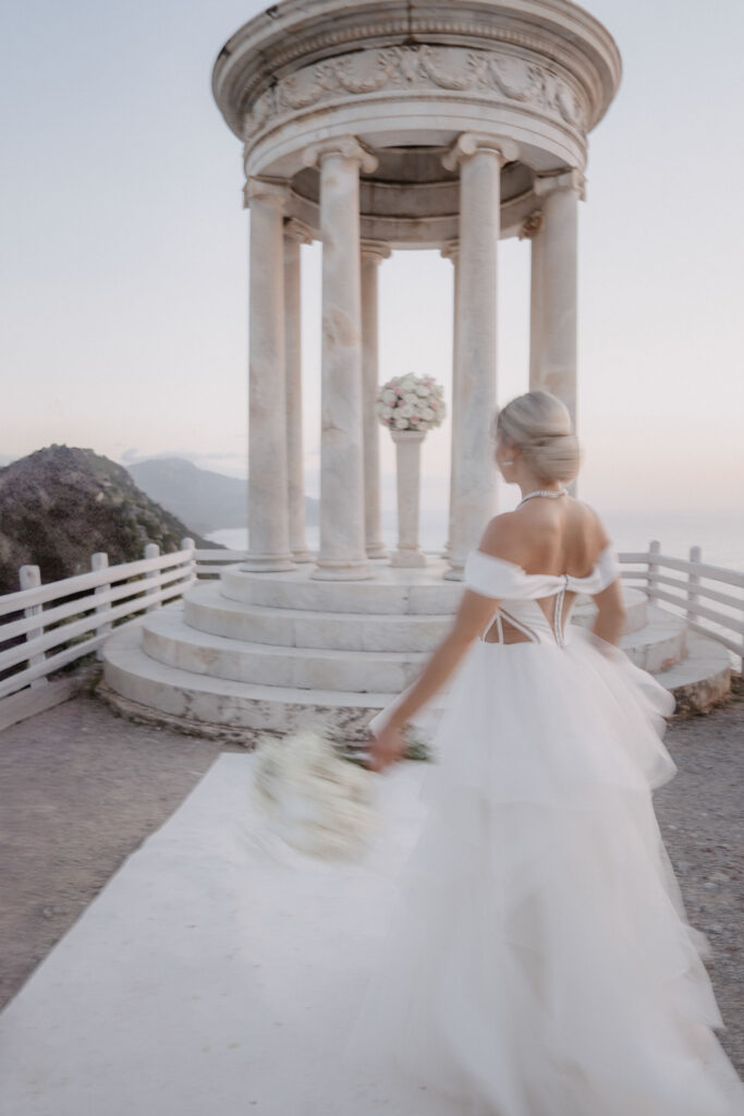 Wedding in Son Marroig: Getting married over the cliffs of Mallorca, with experience, costs and tips 34 A bride in a white dress walks to a marble pavilion with a bouquet of flowers in her hand, with the mountains in the background - perfect for wedding photography abroad.