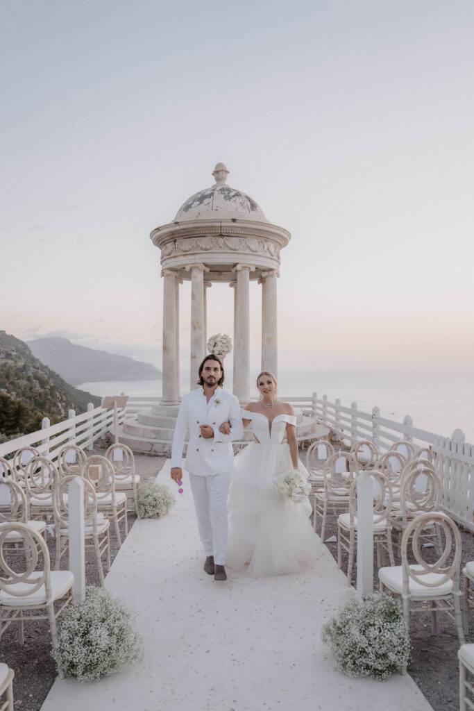 Wedding in Son Marroig: Getting married over the cliffs of Mallorca, with experience, costs and tips 36 A bride and groom walk along an outdoor path that is intended for a wedding at the holiday resort, with a cupola pavilion and sea views in the background – perfect for unforgettable wedding photography abroad.