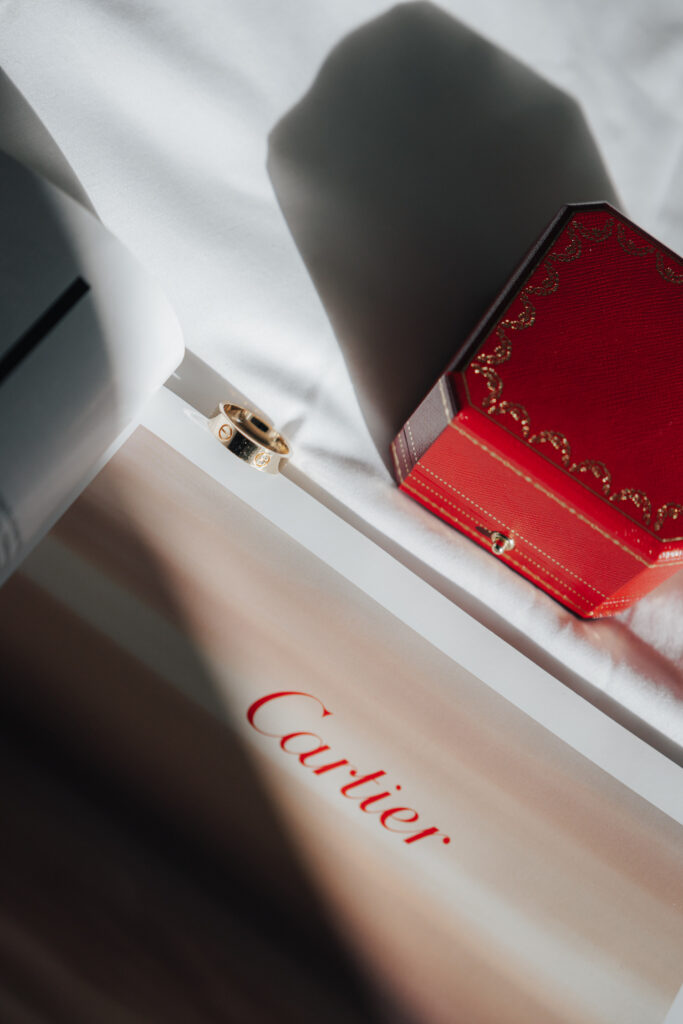A golden ring, a red Cartier box, and a Cartier booklet arranged on a white surface in the sunlight – the perfect inspiration for any wedding photographer or lover of beautiful wedding photography.