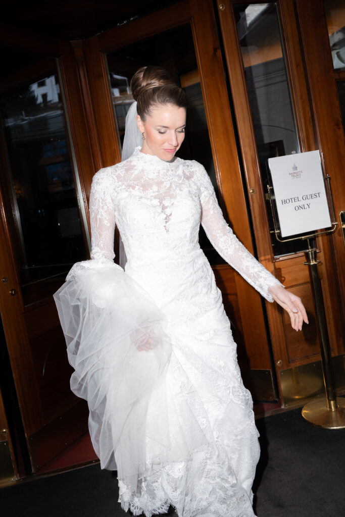 A woman in a long-sleeved white lace wedding dress emerges from a doorway, holding her veil – beautifully captured by a destination wedding photographer for wedding photography abroad.