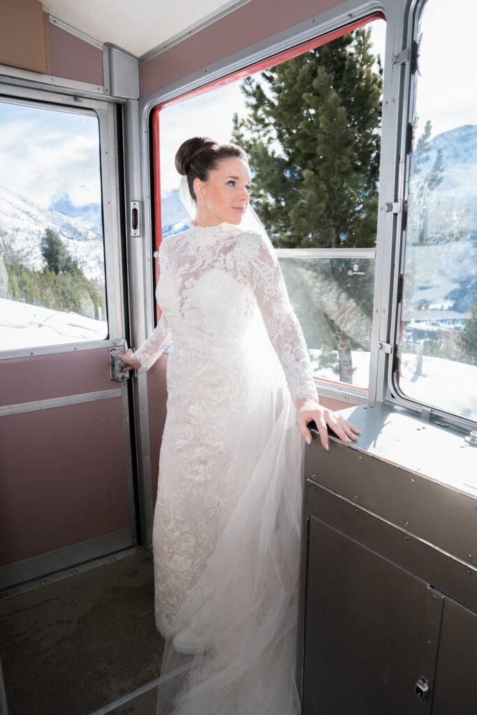 The bride in a lace wedding dress stands in a cable car, against a backdrop of snowy mountains and trees – a dreamy scene perfect for destination wedding photography and elopements.