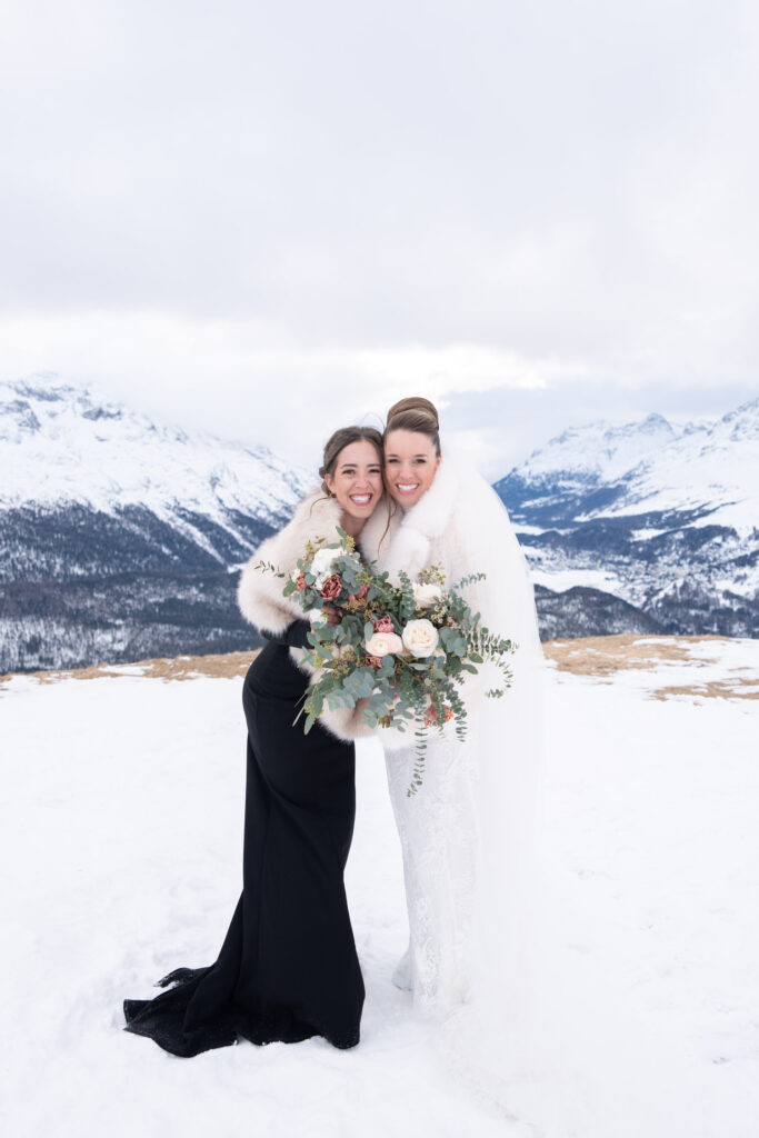 Two women in elegant dresses smile and pose with a bouquet of flowers against a snowy mountain backdrop – perfect overseas wedding photography.
