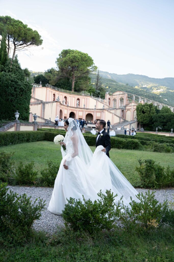 Heiraten am Gardasee: Locations, Kosten und ehrliche Erfahrungen aus über 15 Hochzeiten 1 Braut und Bräutigam spazieren vor einem großen Gebäude in Italien, umgeben von Grün und Gästen im Hintergrund - perfekt für unvergessliche Hochzeitsfotografie bei Auslandshochzeiten.