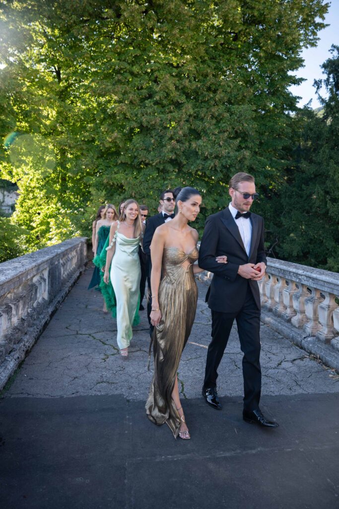 Heiraten am Gardasee: Locations, Kosten und ehrliche Erfahrungen aus über 15 Hochzeiten 14 Eine Gruppe formell gekleideter Menschen geht gemeinsam auf einer Steinbrücke im Freien, umgeben von Grün, und fängt die Eleganz von Auslandshochzeiten und den Charme der Hochzeitsfotografie in Italien ein.