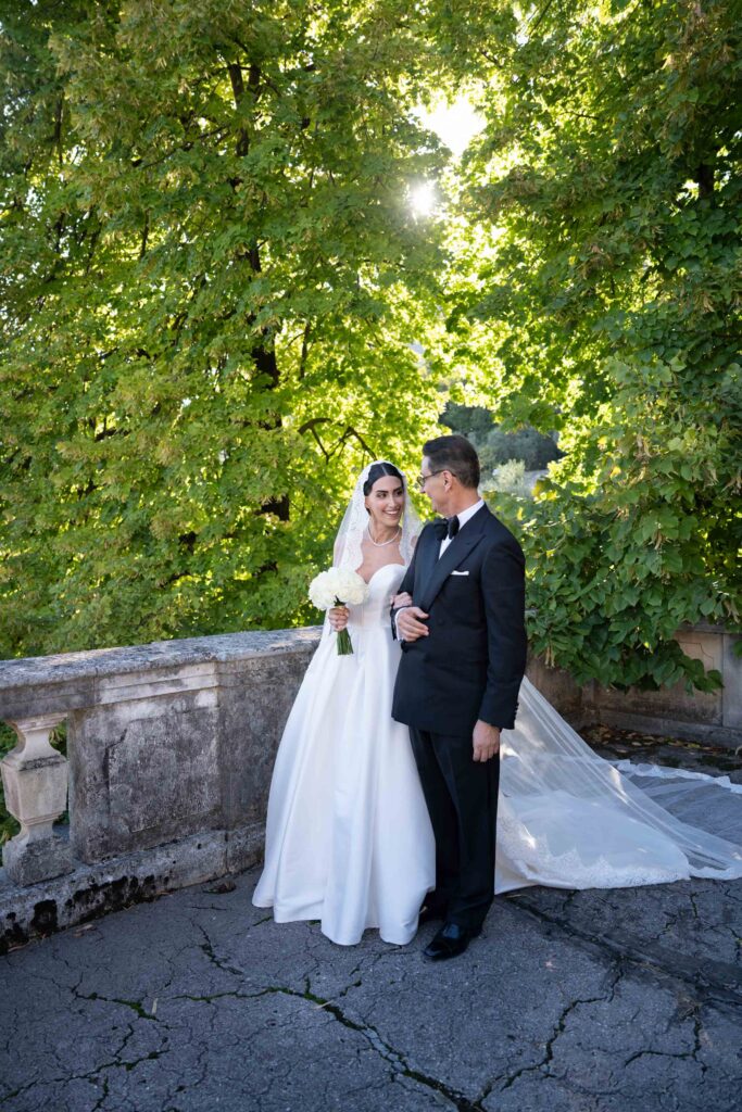 Heiraten am Gardasee: Locations, Kosten und ehrliche Erfahrungen aus über 15 Hochzeiten 15 Braut und Bräutigam stehen gemeinsam im Freien an einem steinernen Geländer, umgeben von üppig grünen Bäumen und Sonnenlicht - perfekte Momente für unvergessliche Hochzeitsfotografie bei Ihrer Destination Wedding.