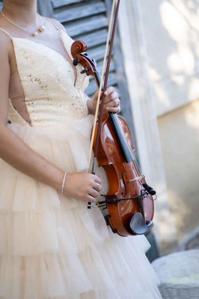 Heiraten am Gardasee: Locations, Kosten und ehrliche Erfahrungen aus über 15 Hochzeiten 17 Eine Person in einem cremefarbenen Paillettenkleid, die eine Geige und einen Bogen in der Hand hält, steht im Freien vor einem Fenster mit Fensterläden - die perfekte Inspiration für eine elegante italienische Hochzeit oder eine romantische Feier auf Mallorca.