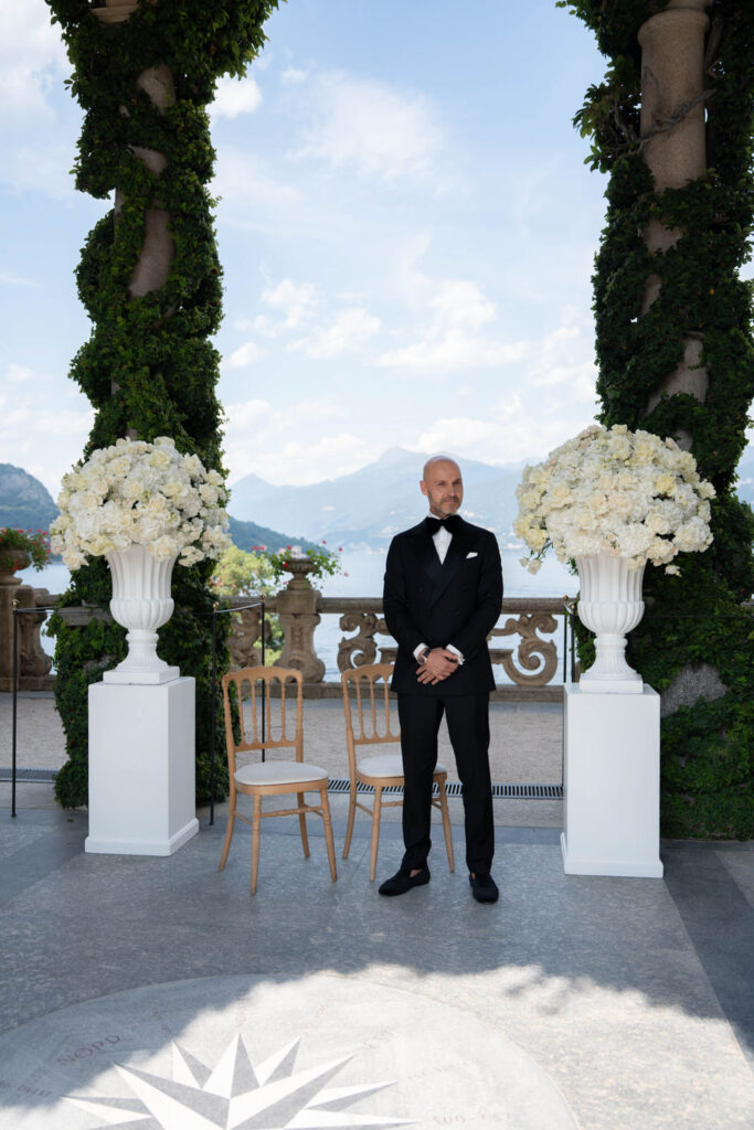 Villa del Balbianello Hochzeit: Kosten, Ablauf und die ikonischste Location am Comer See 17 In Villa del Balbianello, a man in a black tuxedo stands between two large white flower arrangements.