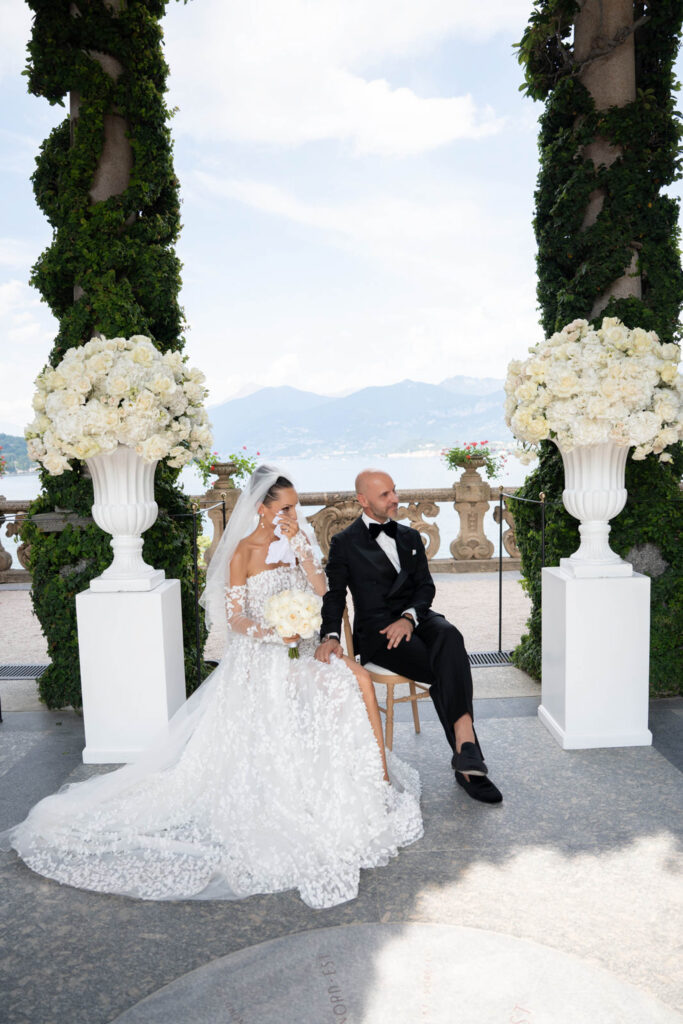 Villa del Balbianello Hochzeit: Kosten, Ablauf und die ikonischste Location am Comer See 18 A bride in a lace dress and a groom in a tuxedo sit together in Villa del Balbianello.