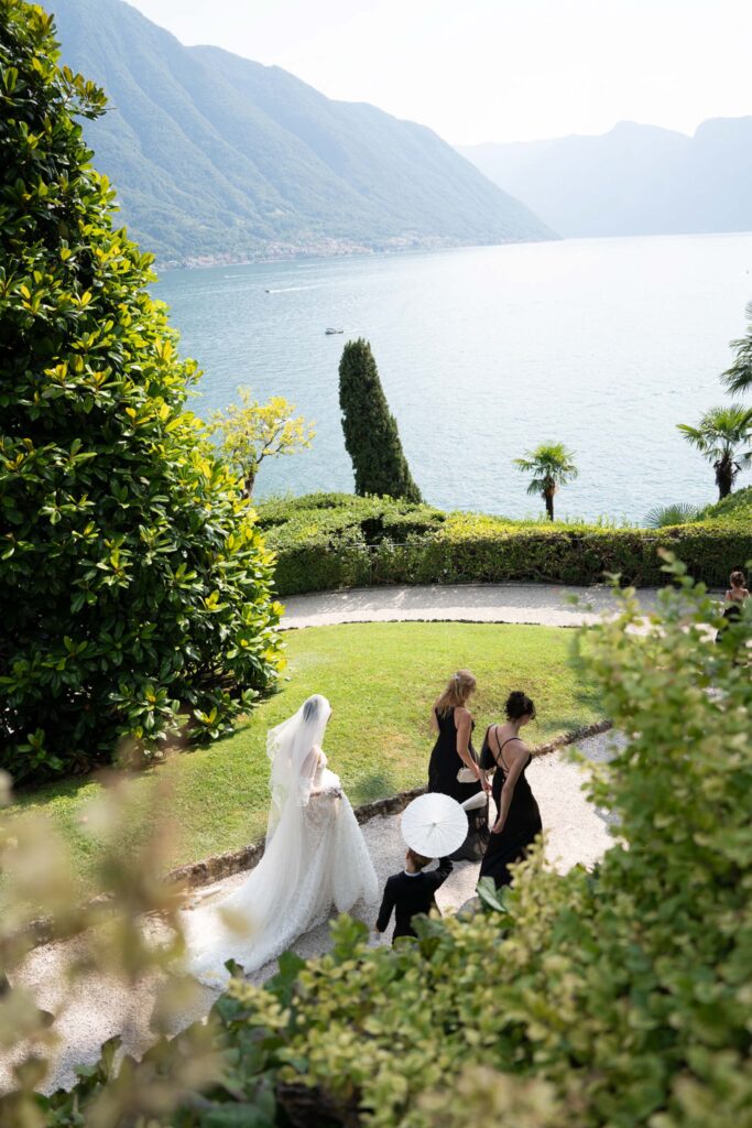 Villa del Balbianello Hochzeit: Kosten, Ablauf und die ikonischste Location am Comer See 55 A bride and two bridesmaids are walking along a garden path in Villa del Balbianello overlooking a lake.