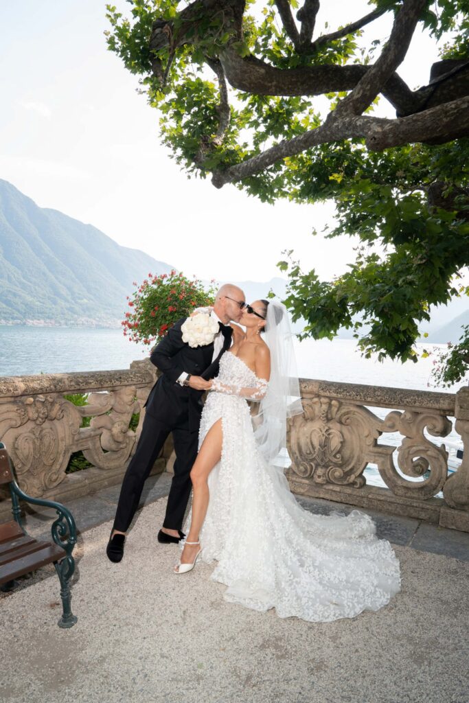Villa del Balbianello Hochzeit: Kosten, Ablauf und die ikonischste Location am Comer See 53 A couple in wedding attire kisses on a balcony of Villa del Balbianello overlooking a lake.