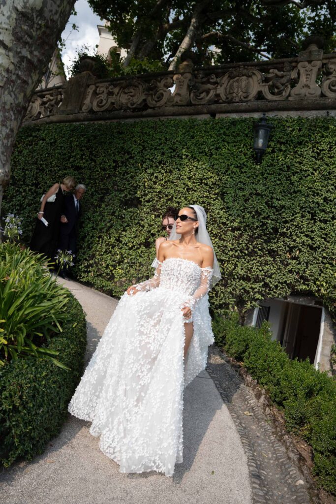 Villa del Balbianello Hochzeit: Kosten, Ablauf und die ikonischste Location am Comer See 51 A bride in a dress with floral appliqués and sunglasses walks along a garden path in Villa del Balbianello.