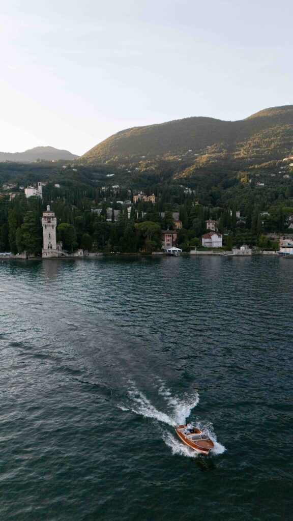 A small wooden boat sails across a lake, with hills and houses along the wooded shore visible in the background – an idyllic setting perfect for destination weddings or wedding photography.