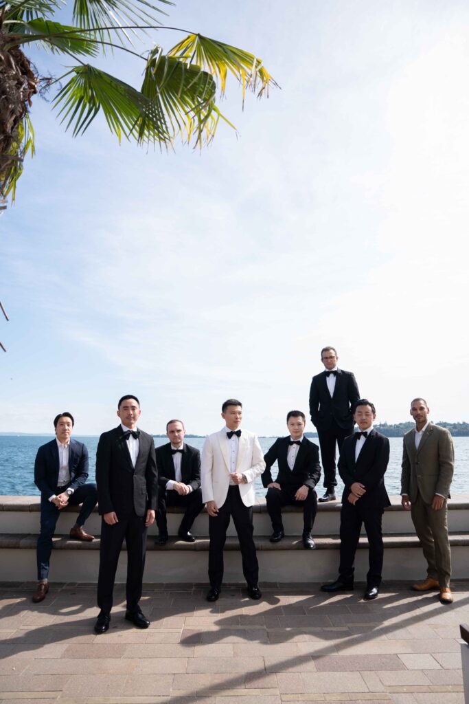 Seven men in formal suits pose outdoors by the water, with a palm tree and blue sky in the background – capturing the spirit of destination wedding photography at a stylish overseas wedding.